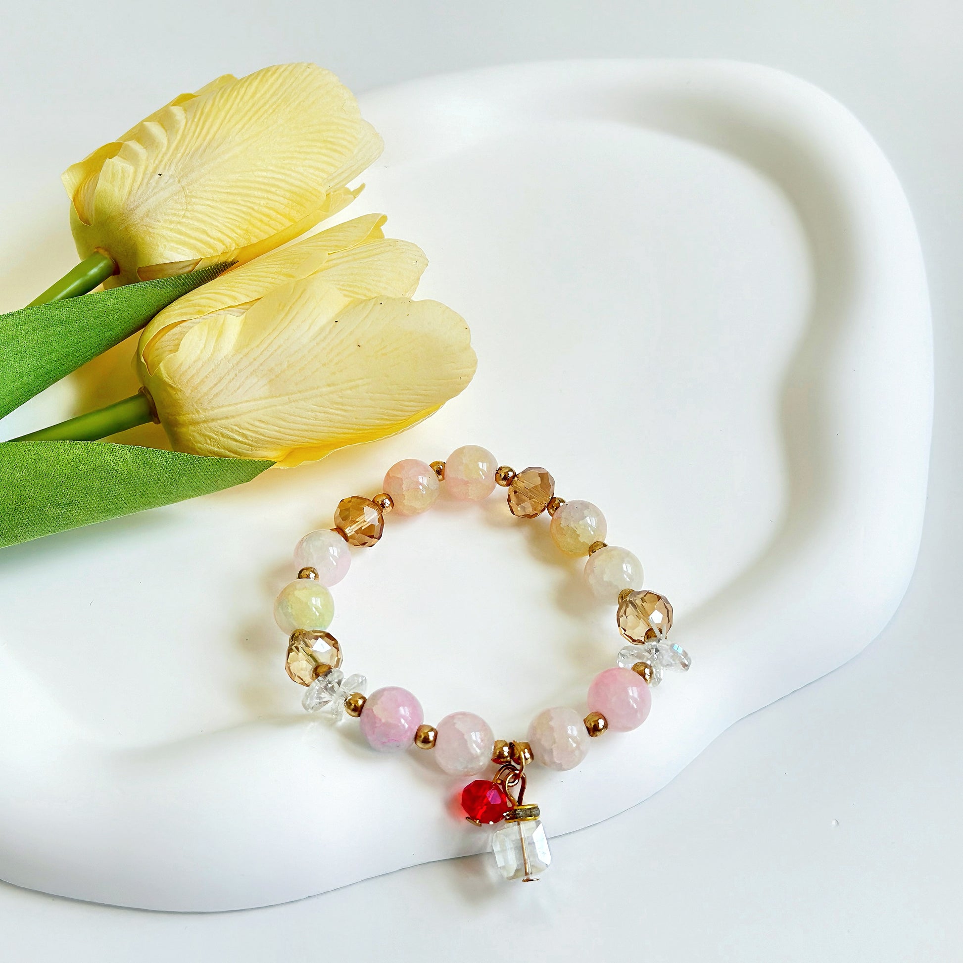 A stunning simple bracelet with light colored beads and bronze accents, along with a simple glass present charm, sitting next to some yellow flowers.