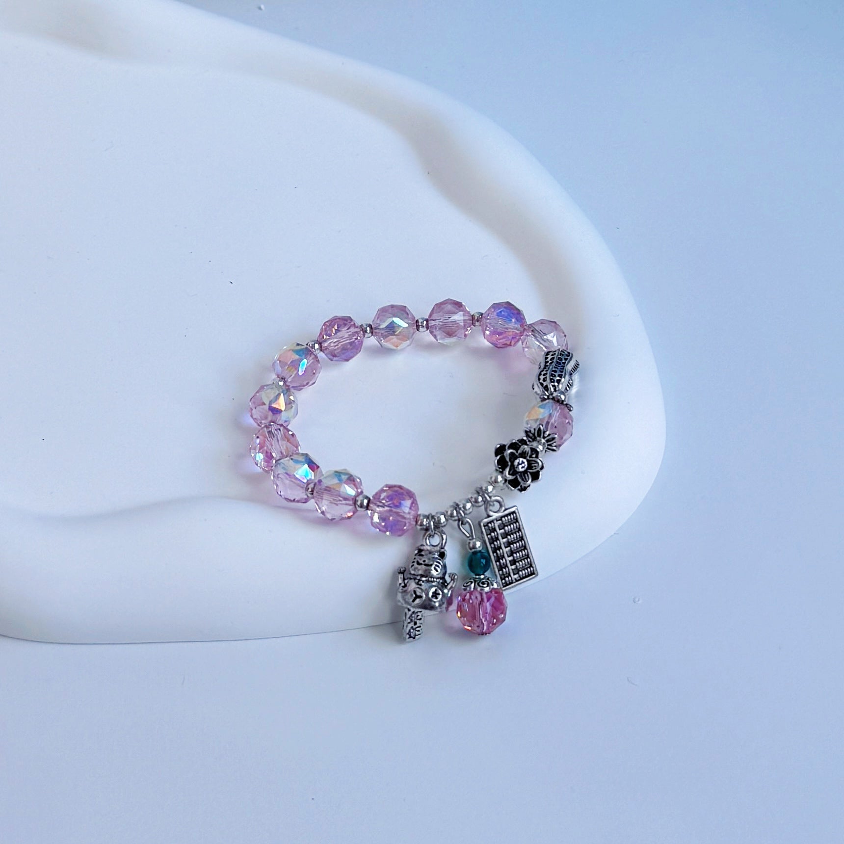 Rose quartz beaded bracelet with charms symbolizing good luck and fortune, including a metal peanut, a metal abacus, a metal lucky cat, and a metal flower.