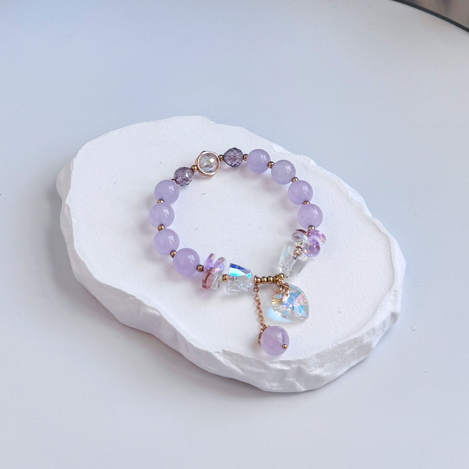 A sophisticated, pretty bracelet with 8 light pink jade beads, each separated by a small gold bead, along with several glass beads, both clear and purple colored, and a dangling clear glass heart charm and last dangling pink jade bead.