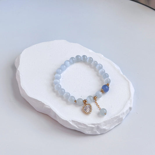 A sophisticated, pretty bracelet with 22 light gray aquamarine beads and a single royal blue bead, clasped with a small dangling glass heart charm and additional gray aquamarine bead.