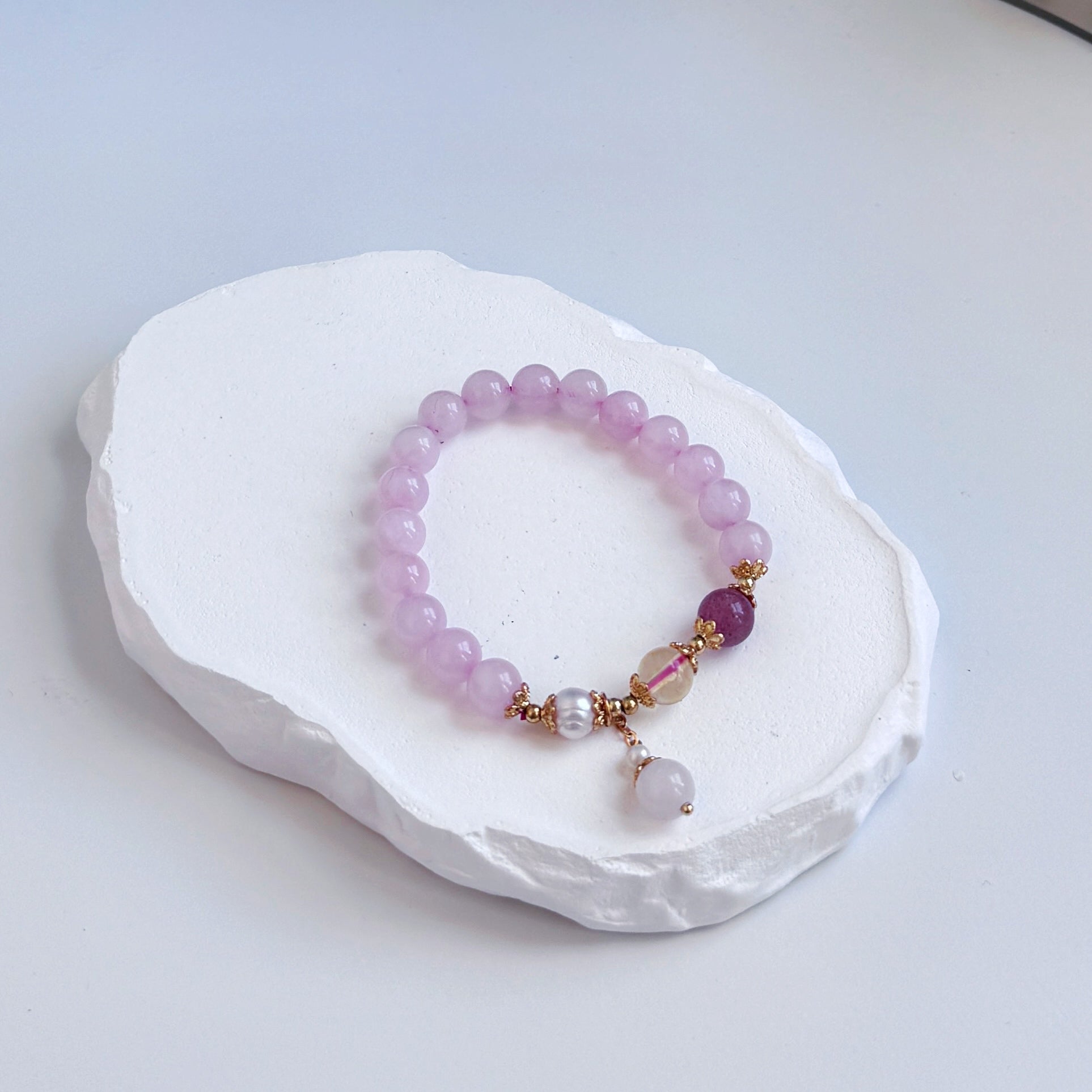 A sophisticated, pretty bracelet with 16 light purple rose quartz beads, along with a pearl, a citrine bead, and a darker purple strawberry quartz bead, all with golden colored accents.