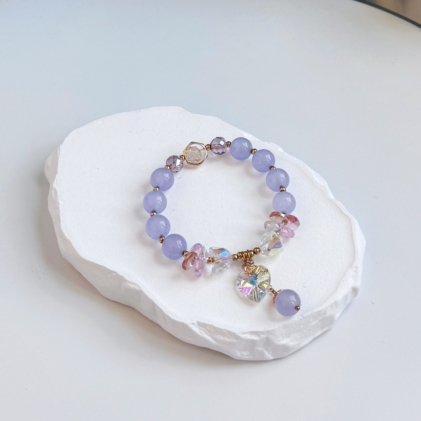 A sophisticated, pretty bracelet with 8 light purple amethyst beads, each separated by a small gold bead, along with several glass beads, both clear and purple colored, and a dangling clear glass heart charm and last dangling amethyst bead.