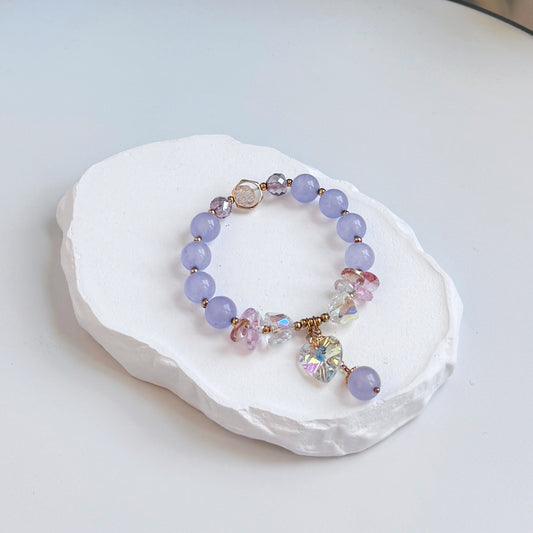 A sophisticated, pretty bracelet with 8 light purple amethyst beads, each separated by a small gold bead, along with several glass beads, both clear and purple colored, and a dangling clear glass heart charm and last dangling amethyst bead.