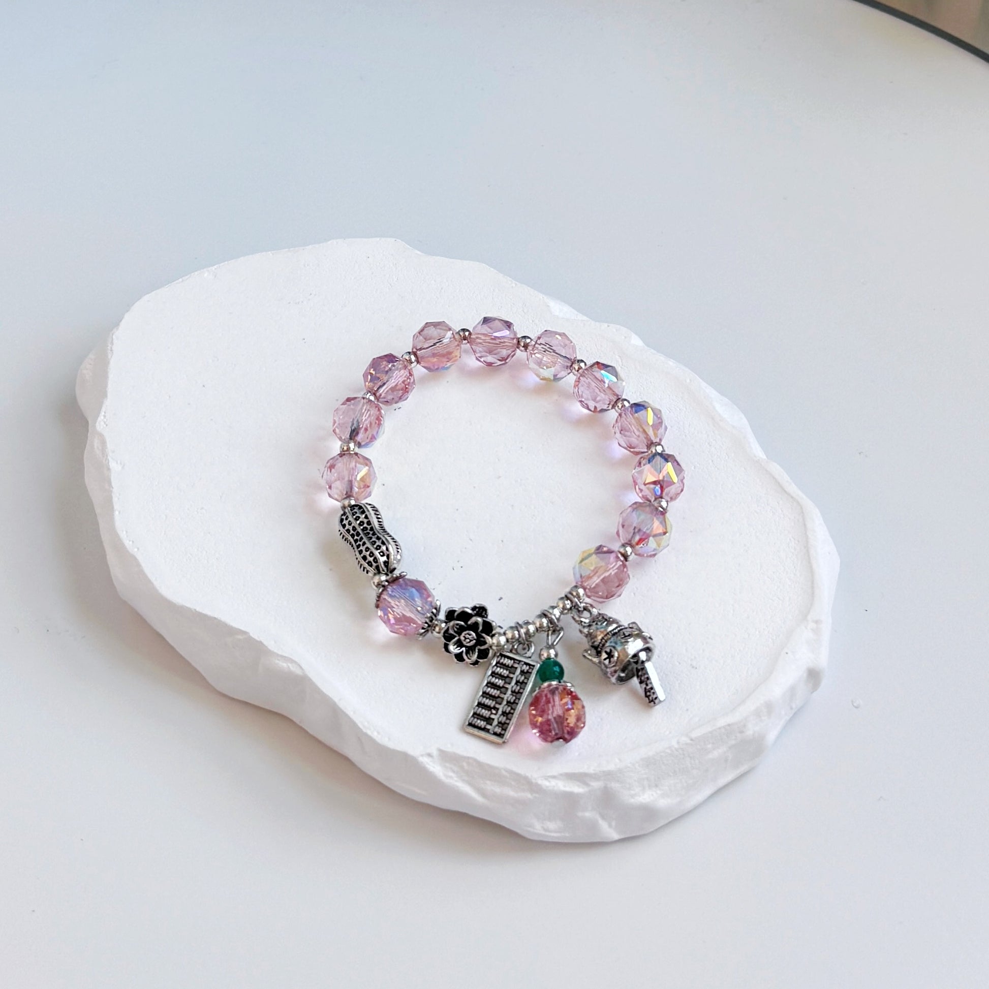 A sophisticated, pretty bracelet with 12 rose quartz beads in a light pink hue, each separated by a small silver colored accent bead, with beads and charms charms symbolizing good luck and fortune, including a metal peanut, a metal abacus, a metal lucky cat, and a metal flower.