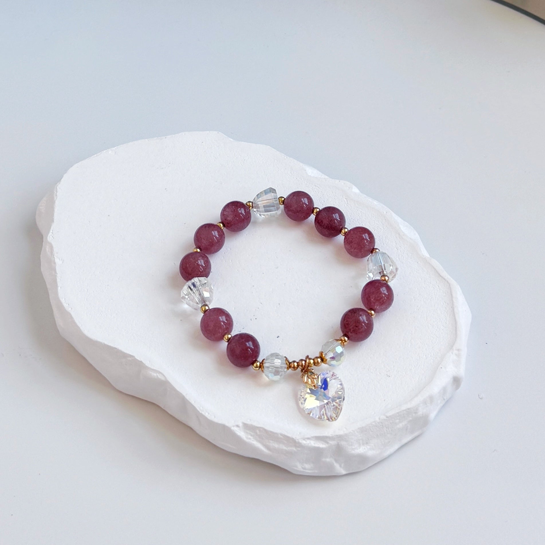 A sophisticated, pretty bracelet with 15 velvet colored strawberry quartz and clear glass beads, each separated by a small gold colored accent bead, and with a small dangling heart charm made of Swarovski crystal.