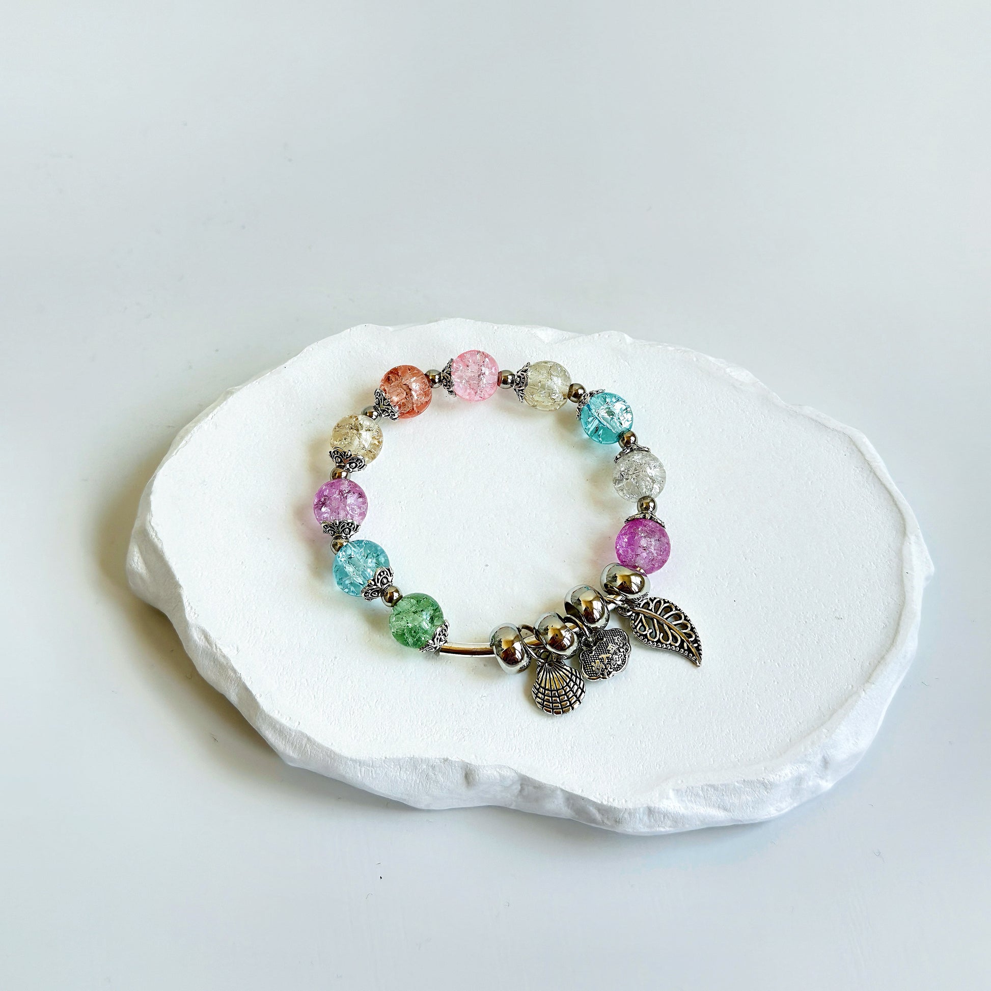 A simple, cute bracelet with 10 glass beads of various light colors, including light blue, light green, light pink, light purple, and crystal white, each separated by a small metal bead, tied with several metal charms, including a clam and leaf.