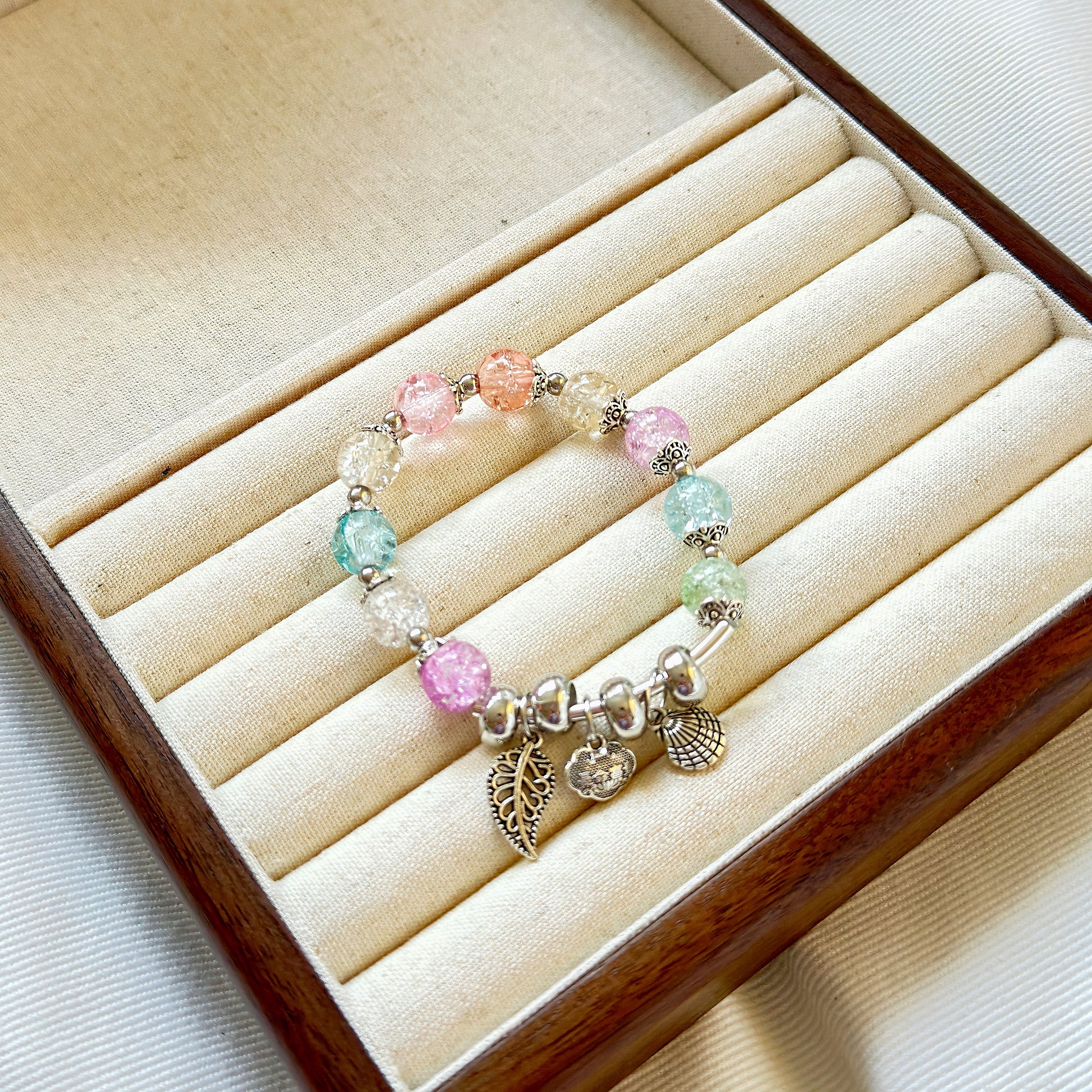 A pretty, delicate, colorful bracelet with colors and charms that resemble a mermaid's under-the-sea paradise, sitting in a wooden jewelry tray.