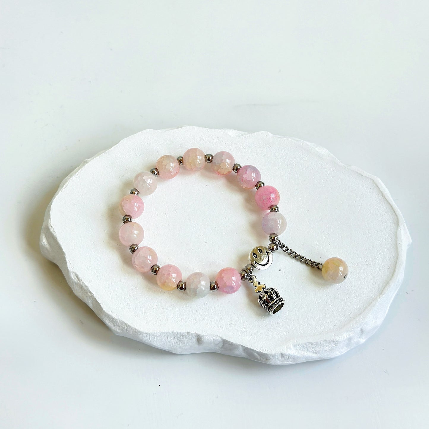 A simple, cute bracelet with 13 light pink glass beads each separated by a small metal bead, clasped with a metal smiley face bead, a metal crown charm, and a dangling last glass bead.