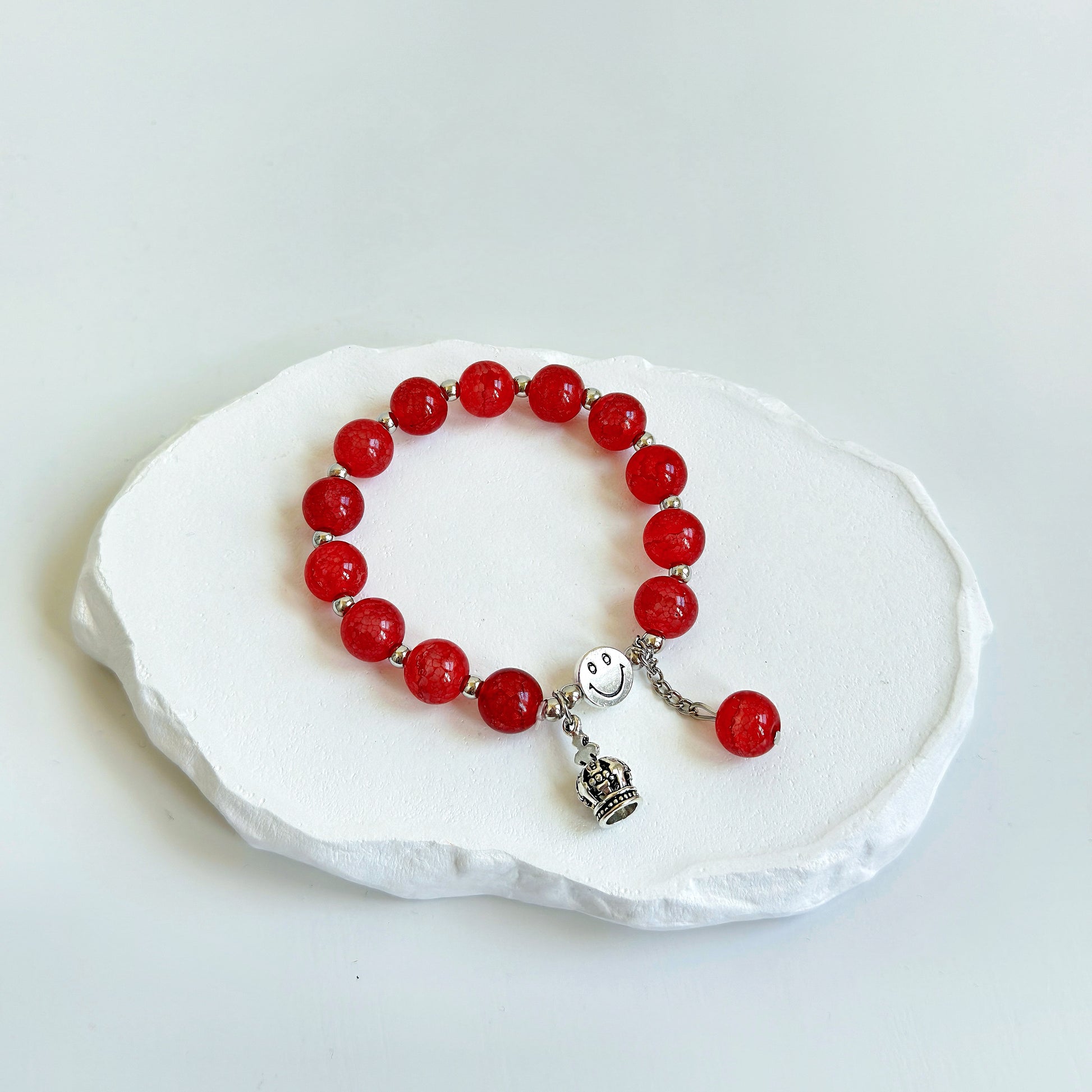 A simple, cute bracelet with 13 crimson-red glass beads each separated by a small metal bead, clasped with a metal smiley face bead, a metal crown charm, and a dangling last glass bead.