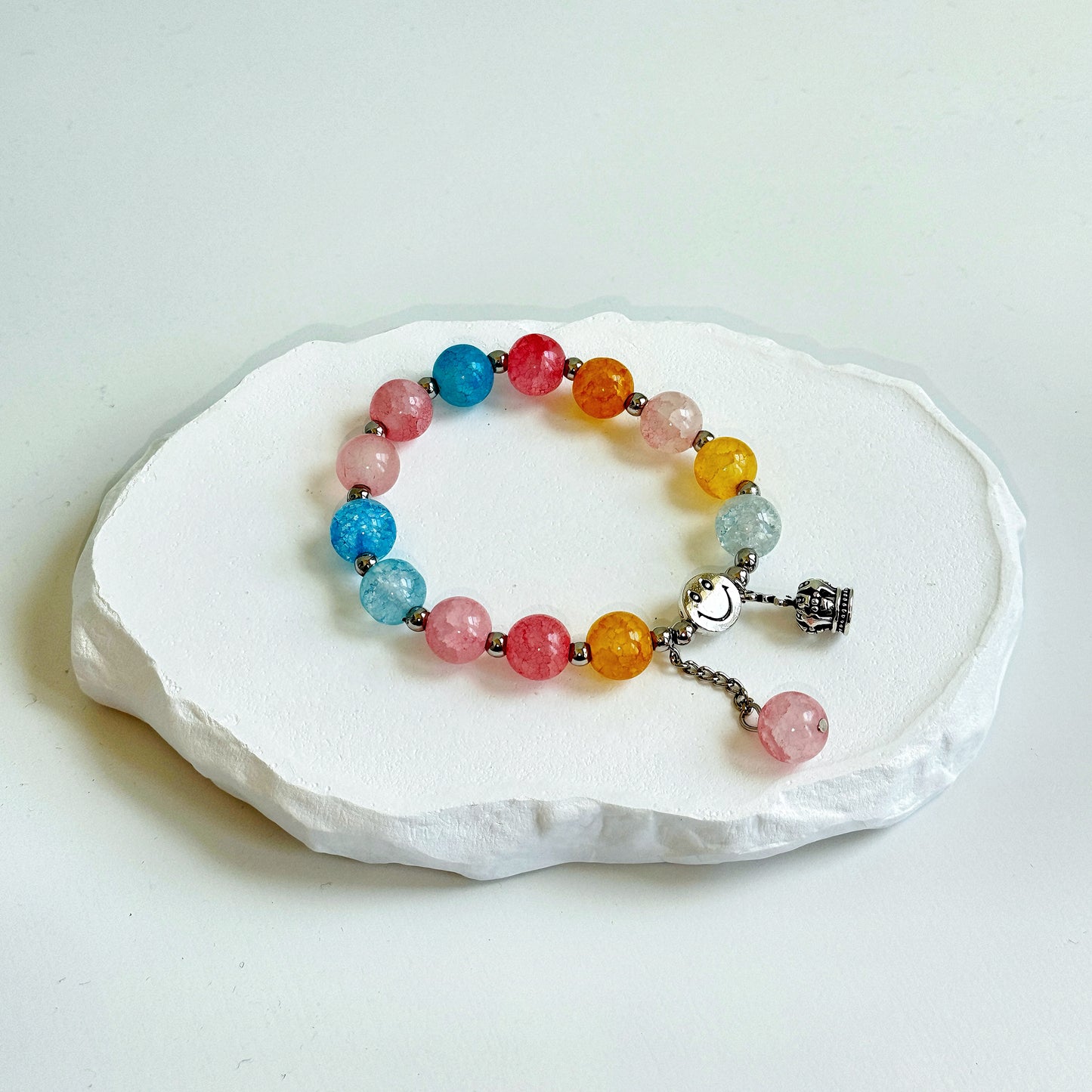 A simple, cute bracelet with 13 glass beads, with colors such as light blue, light pink, gold, and crystal white, each separated by a small metal bead, clasped with a metal smiley face bead, a metal crown charm, and a dangling last glass bead.