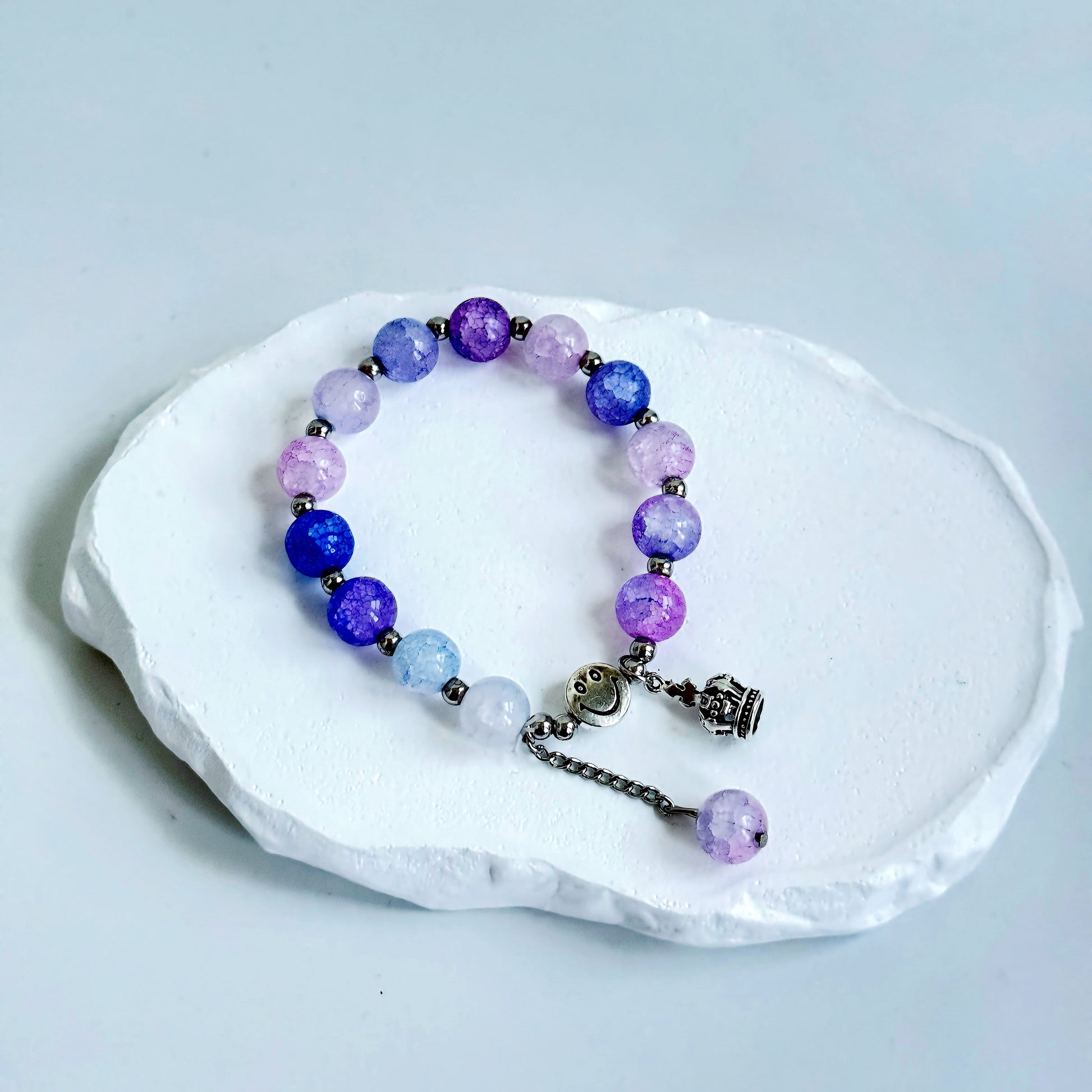 A simple, cute bracelet with 13 lavender and purple glass beads each separated by a small metal bead, clasped with a metal smiley face bead, a metal crown charm, and a dangling last glass bead.