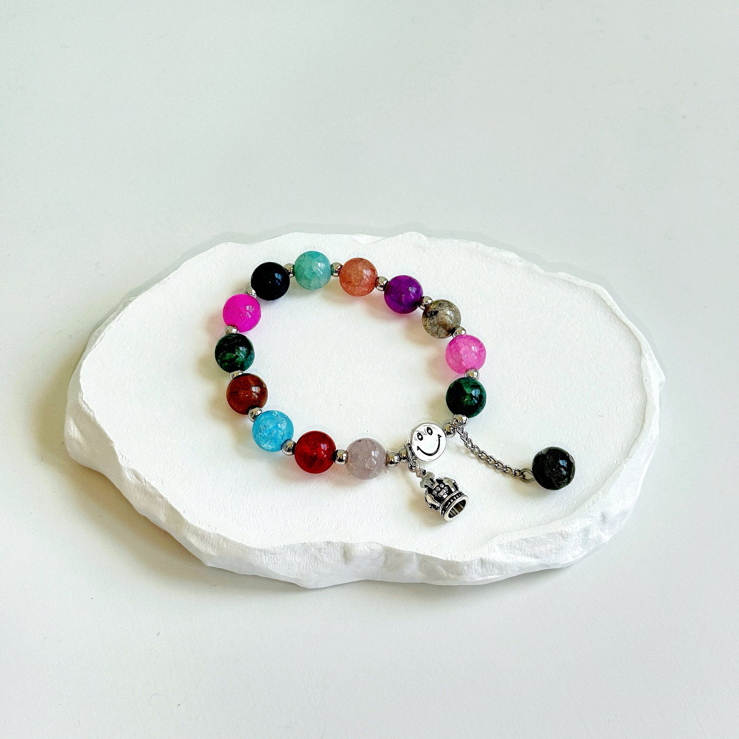 A simple, cute bracelet with 13 glass beads, with a vibrant mix of colors, including pink, green, red, blue, and white, each separated by a small metal bead, clasped with a metal smiley face bead, a metal crown charm, and a dangling last glass bead.