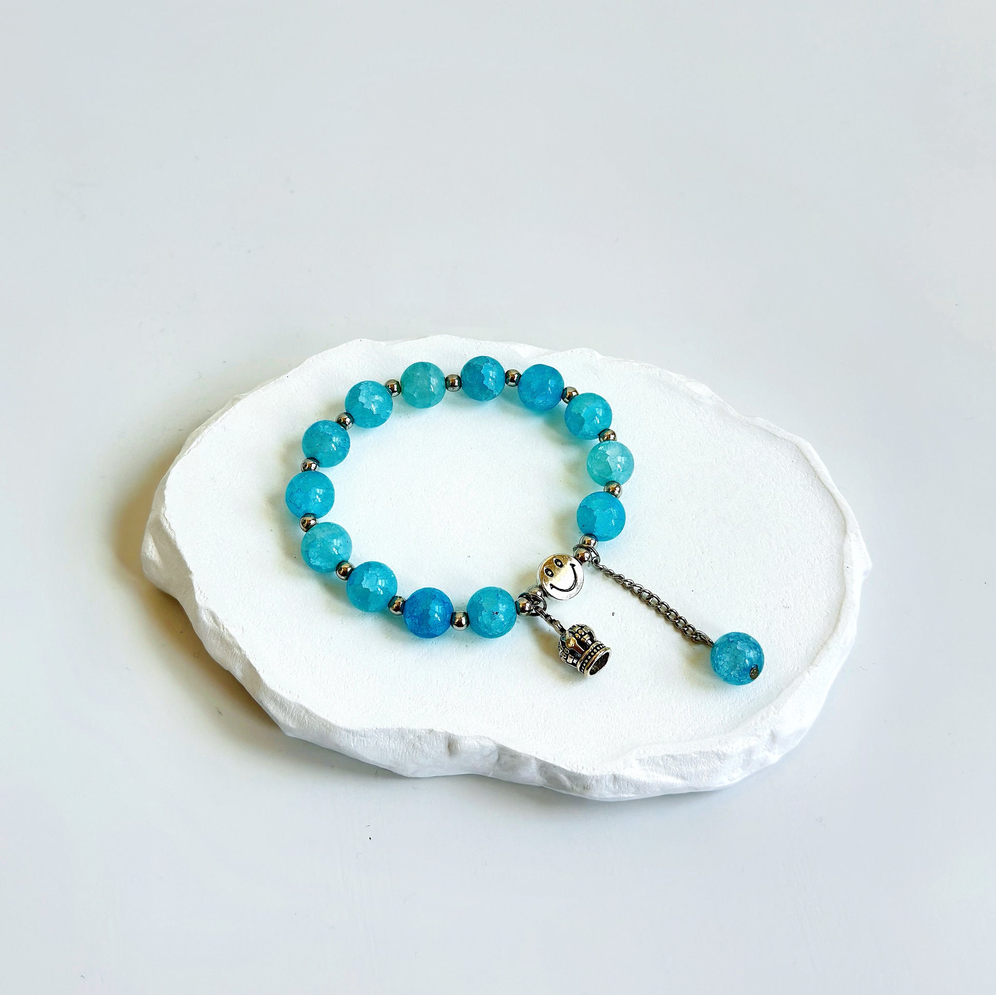 A simple, cute bracelet with 13 light blue glass beads each separated by a small metal bead, clasped with a metal smiley face bead, a metal crown charm, and a dangling last glass bead.