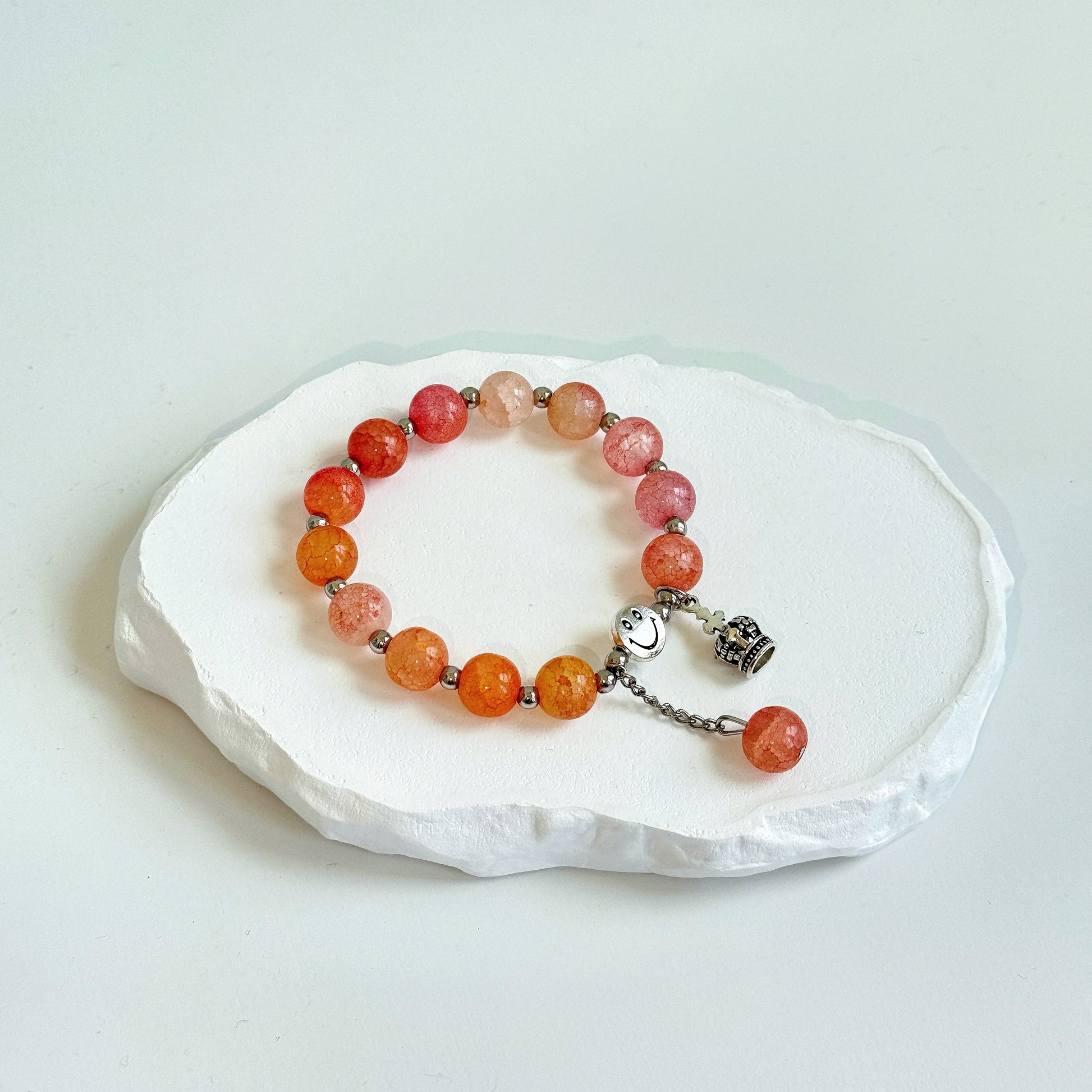 A simple, cute bracelet with 13 glass beads, ranging from colors of vibrant amber, to opaque white, to rosy pink, each separated by a small metal bead, clasped with a metal smiley face bead, a metal crown charm, and a dangling last glass bead.