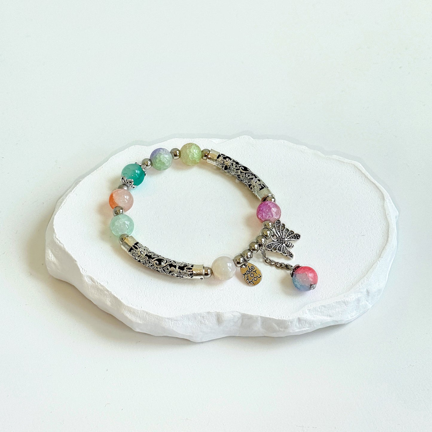 A simple, cute bracelet with 7 crystal glass beads of various colors each separated by a small metal bead, along with several long metal beads, intricately etched with an ornate design, tied with a metal butterfly charm and a dangling last glass bead.